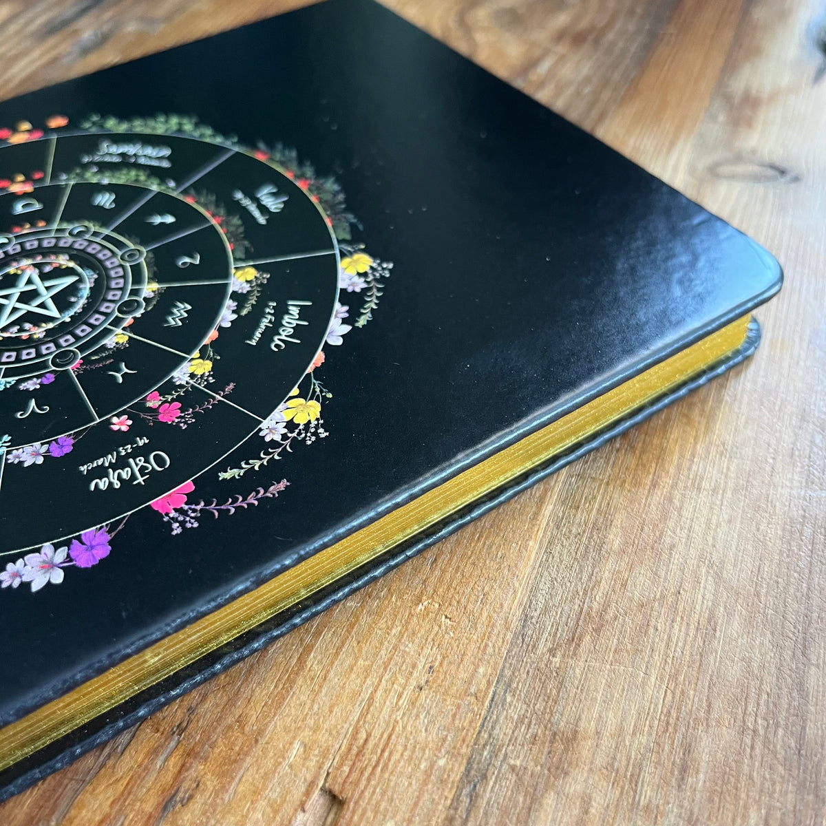A black hardcover journal featuring a detailed circular astrological wheel design with colorful floral accents, resting on a rustic wooden surface.