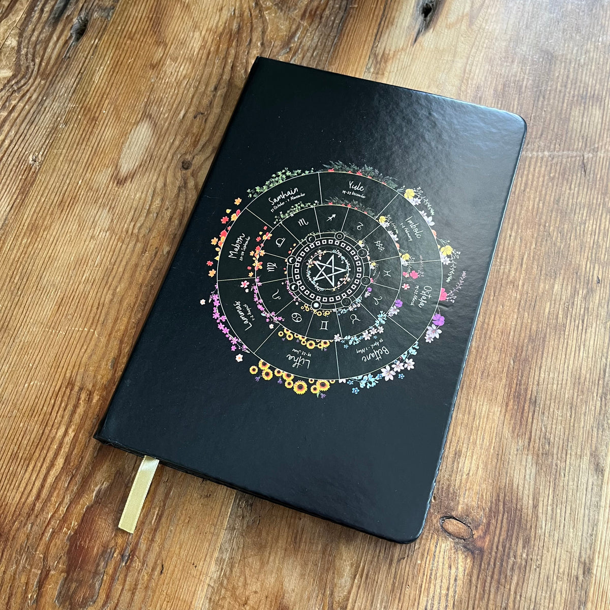 A black hardcover journal featuring a colorful zodiac wheel of the year design on the cover, resting on a rustic wooden table with a gold ribbon bookmark visible.