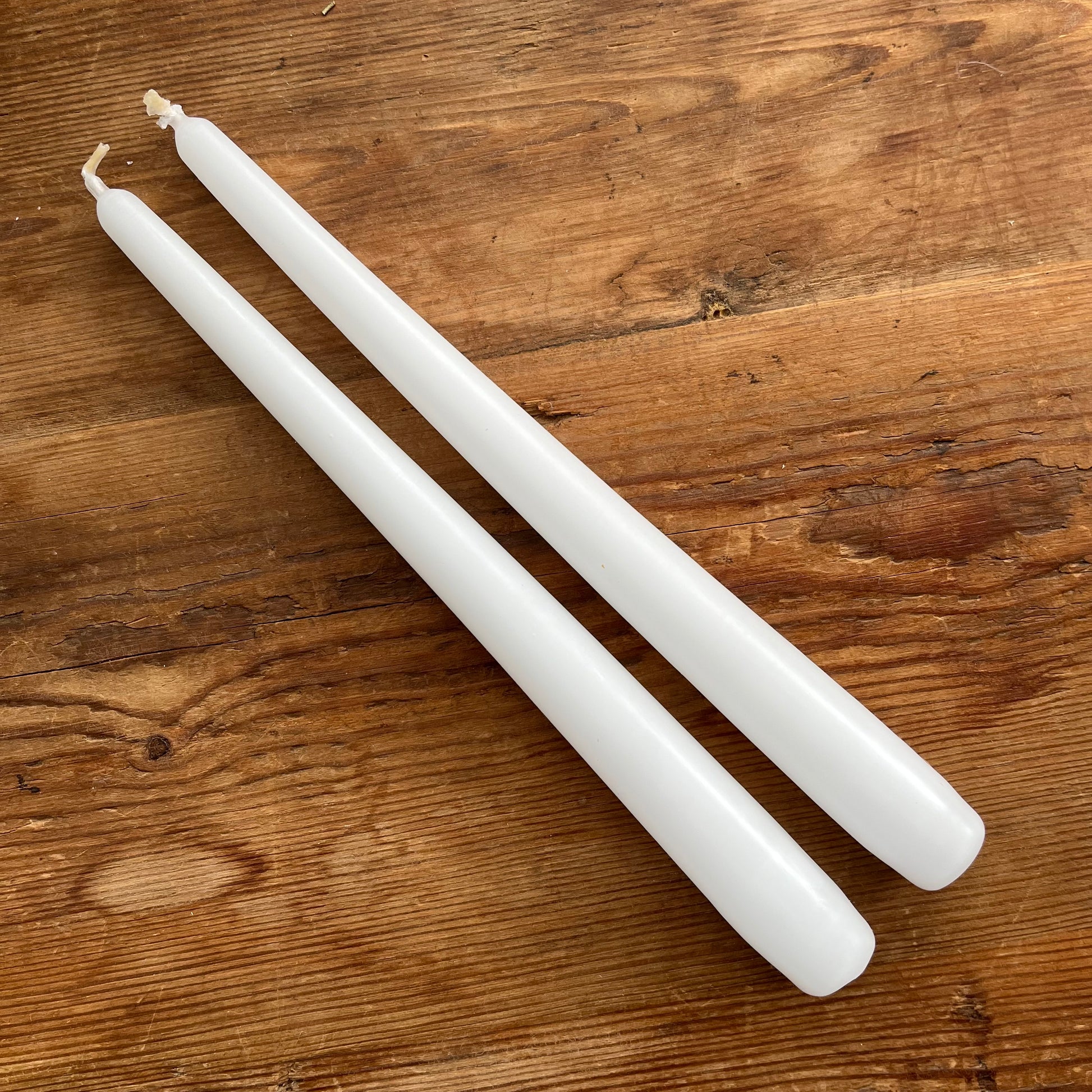 Two white taper candles placed diagonally on a rustic wooden surface.