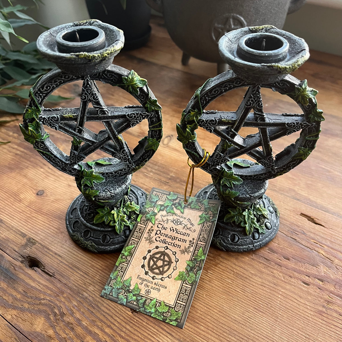 Two dark metal pentacle candle holders with green leaf accents sit on a wooden table, with a printed card reading 'The Wiccan Herbaglum Collection' placed in front.