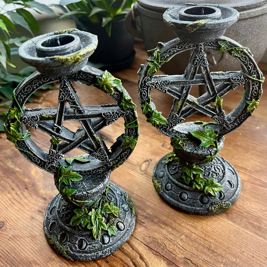 Two dark gray metal pentagram candle holders with green ivy accents, displayed on a wooden tabletop with potted plants in the background.