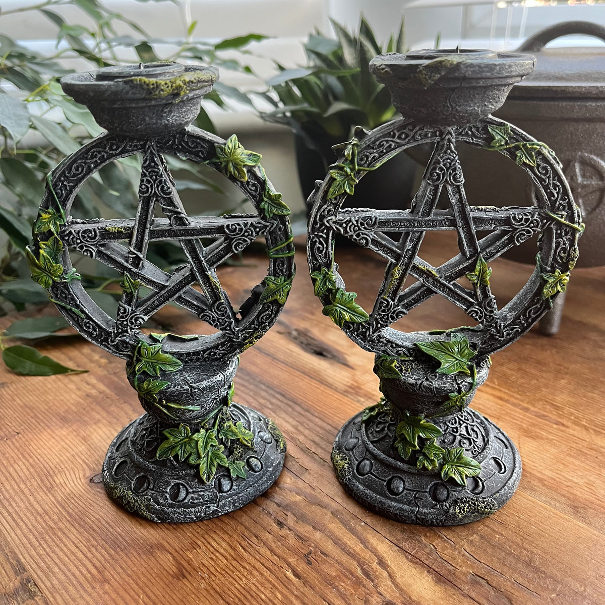 Two dark gray gothic-style metal candle holders shaped like interlocking pentagrams, decorated with faux ivy and moss, displayed on a wooden table with plants in the background.