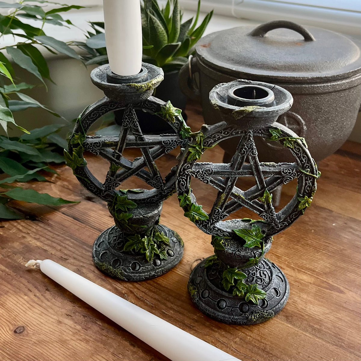 Two dark metal pentagram candle holders with faux ivy details, shown on a wooden table with white candles in a home setting.