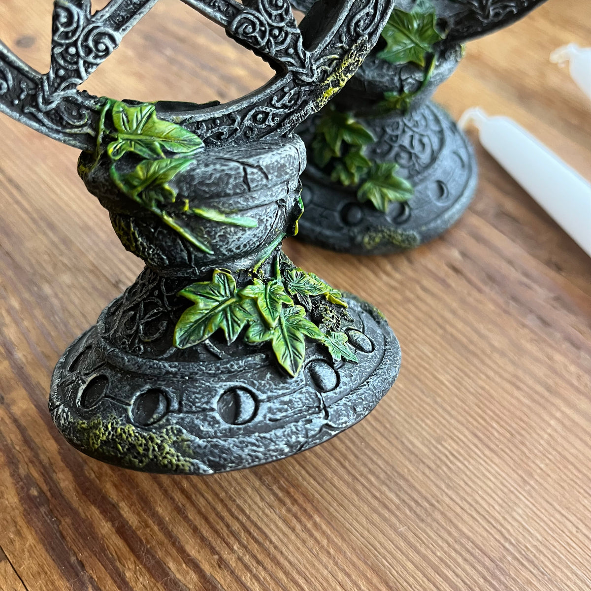 A close-up of a decorative black metal candle holder with green ivy vine details, set on a wooden tabletop next to a white taper candle.