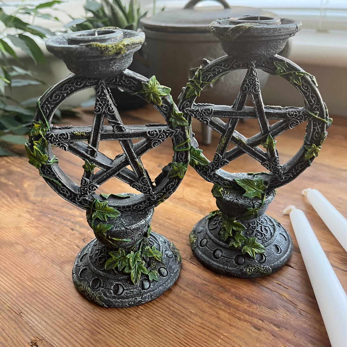Two dark gray metal pentacle candle holders with green ivy accents, displayed on a wooden table next to white taper candles.