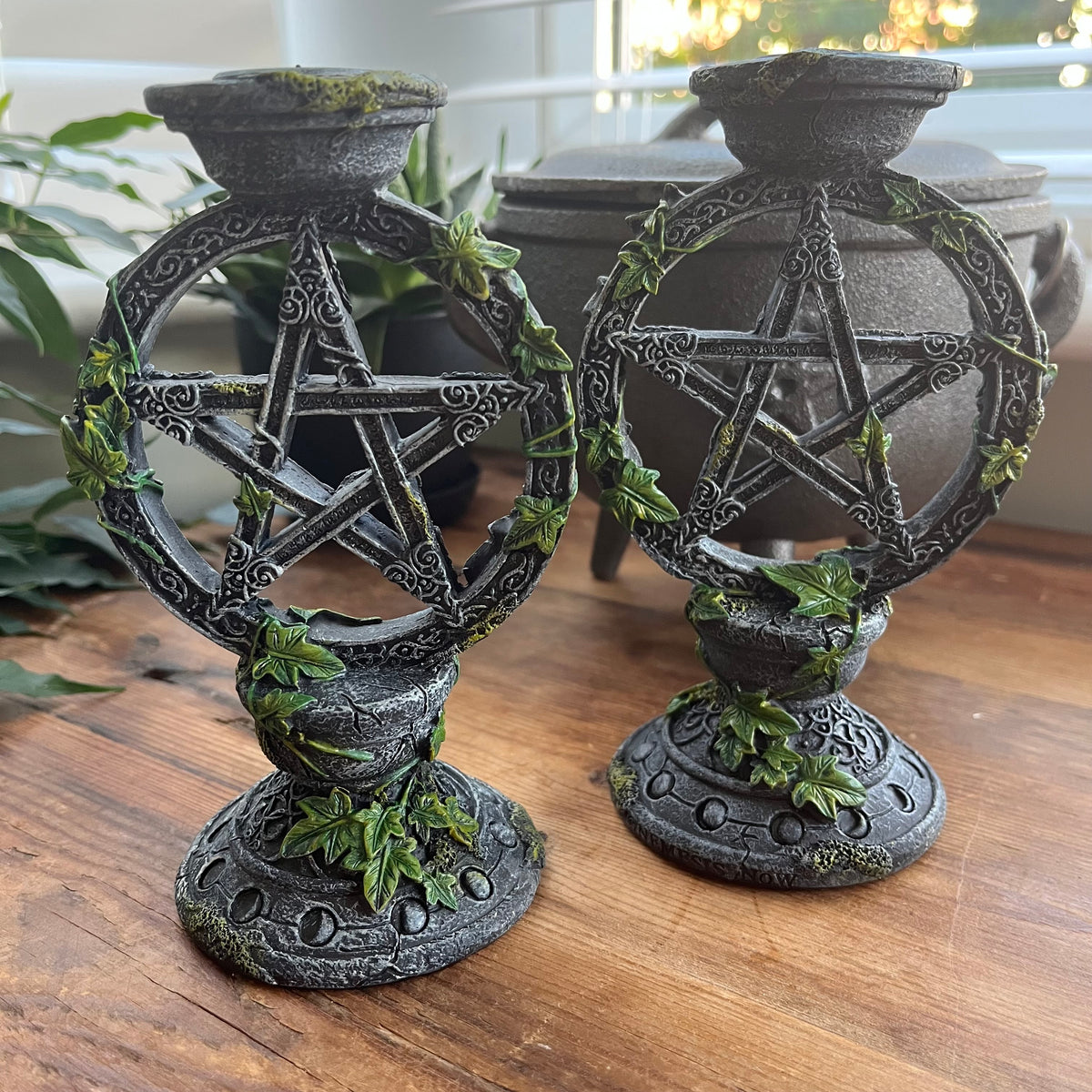 Two dark gray cast-iron-style pentacle candle holders with green ivy accents, displayed on a wooden table with a blurred background.