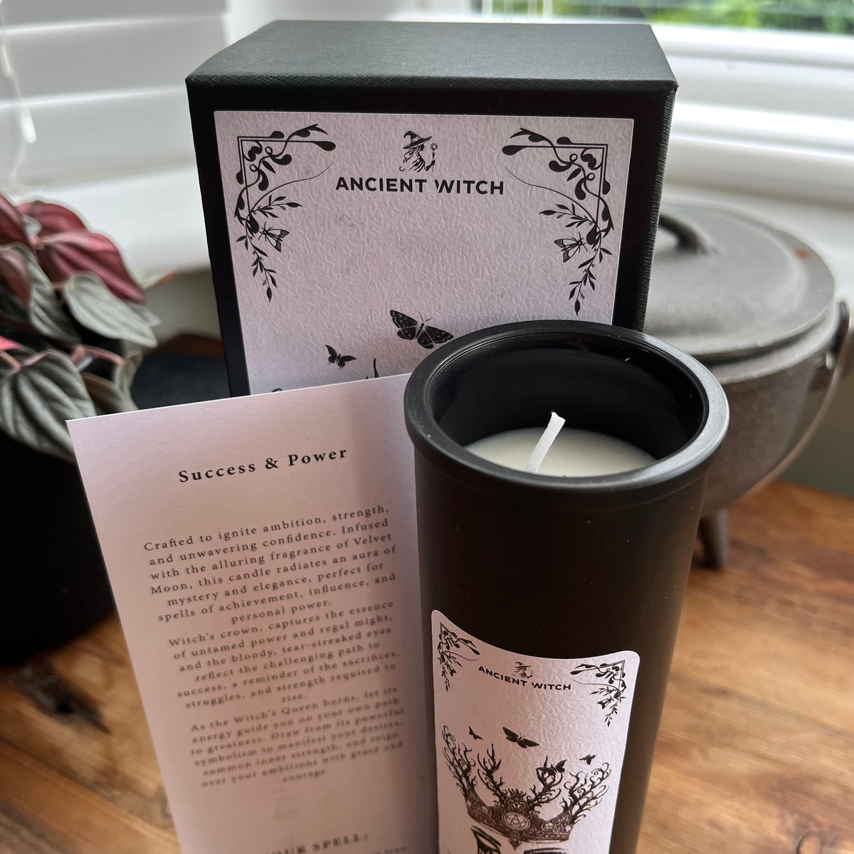 A black candle jar with a white label reading 'ANCIENT WITCH' stands in front of its matching black and white box, with a printed card beside it on a wooden surface near a window.