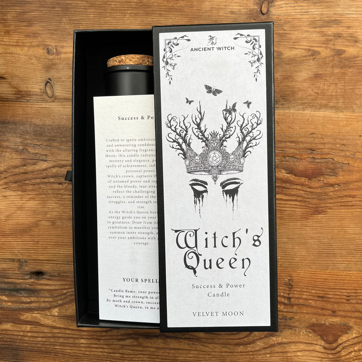 A Witch's Queen candle by Ancient Witch in a black presentation box on a wooden surface. The box is open to reveal a white candle with a cork stopper, next to a tall white card featuring black gothic artwork of a queen with antlers and the text 'Witch's Queen' and 'Ancient Witch'.