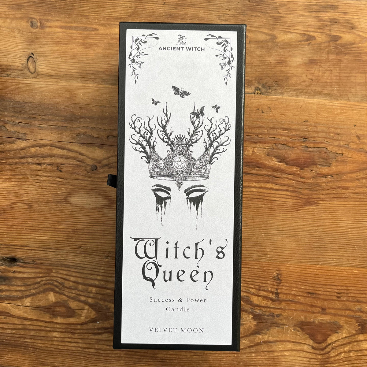 A tall rectangular candle box standing upright on a rustic wooden surface. The box front is white with black gothic-style text reading 'Ancient Witch' at the top and 'Witch's Queen' prominently in the center, plus 'Success & Power Candle' and 'VELVET MOON' at the bottom.
