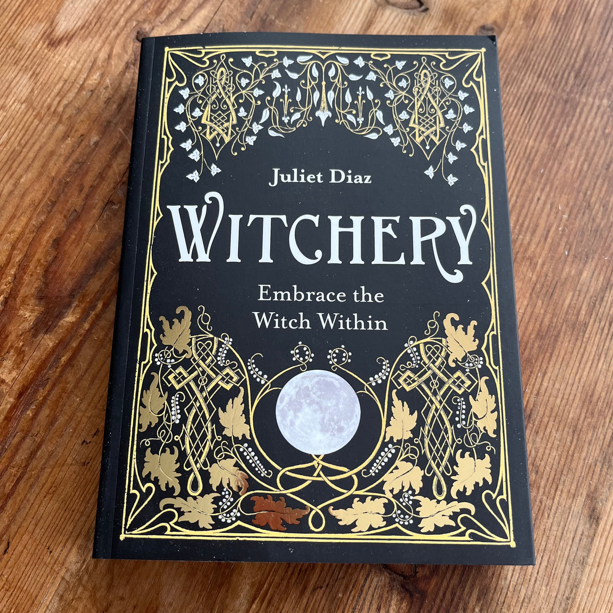 The book 'Witchery' by Juliet Diaz, featuring a black cover with ornate gold and white gothic illustrations and a central moon graphic, photographed flat on a wooden surface.