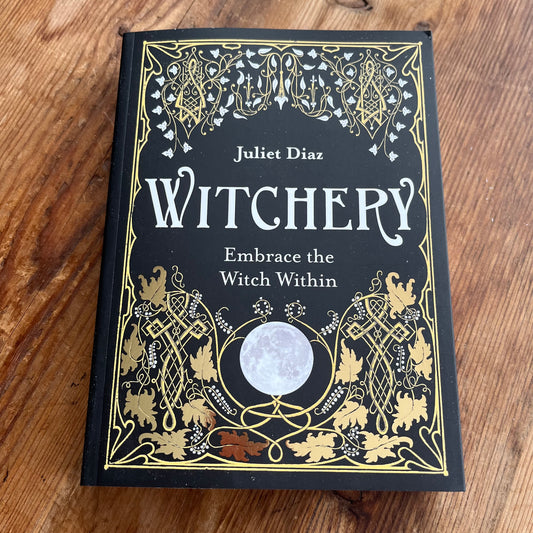 The book 'Witchery' by Juliet Diaz, featuring a black cover with ornate gold and white gothic illustrations and a central moon graphic, photographed flat on a wooden surface.