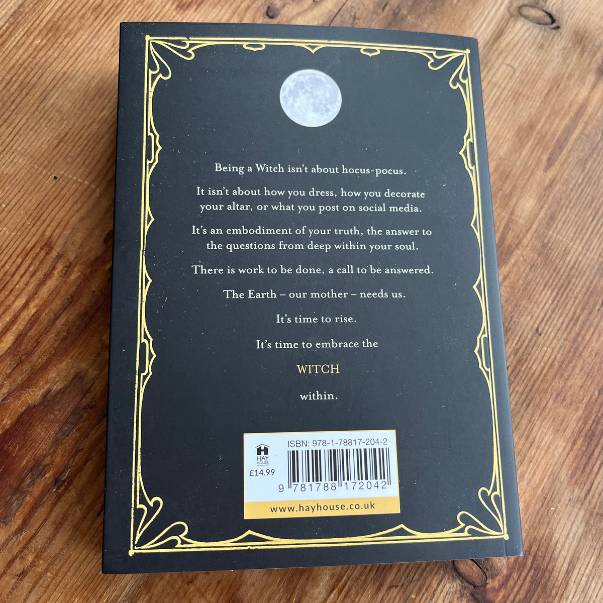 The back cover of a black book titled 'The Witch Within' by Hay House, featuring a gold decorative border, white text, a moon illustration, and a barcode on a wooden surface.