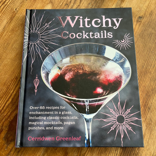 The book 'Witchy Cocktails' by Cerridwen Greenleaf lies flat on a wooden table. The cover is dark gray with pink celestial illustrations and a large image of a martini glass containing a dark red drink.