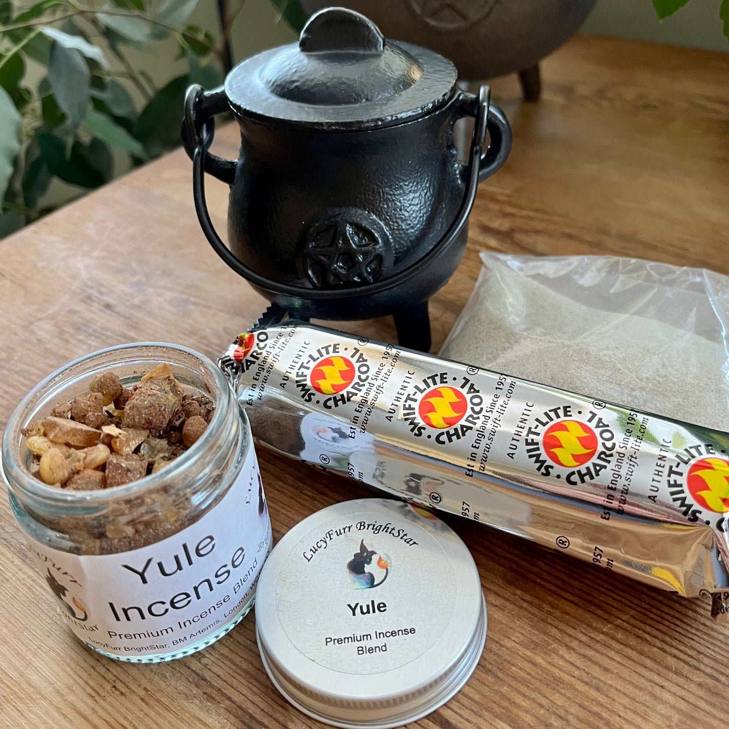 Yule incense jar, Yule incense beads, and a package of Charcoal on a wooden surface with a black cauldron in the background.