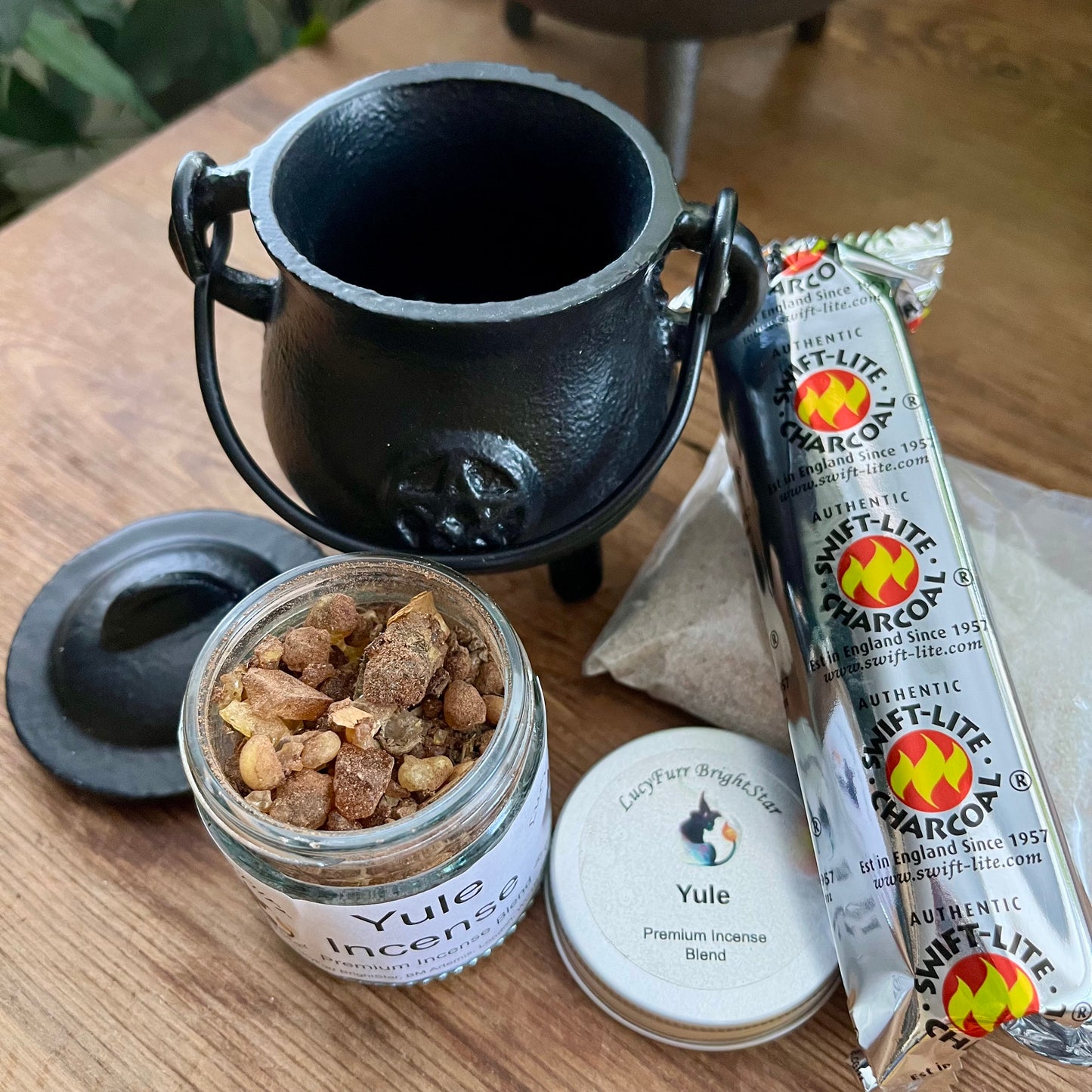 Black cauldron with Yule Incense jar,  and Charcoal product on a wooden surface.