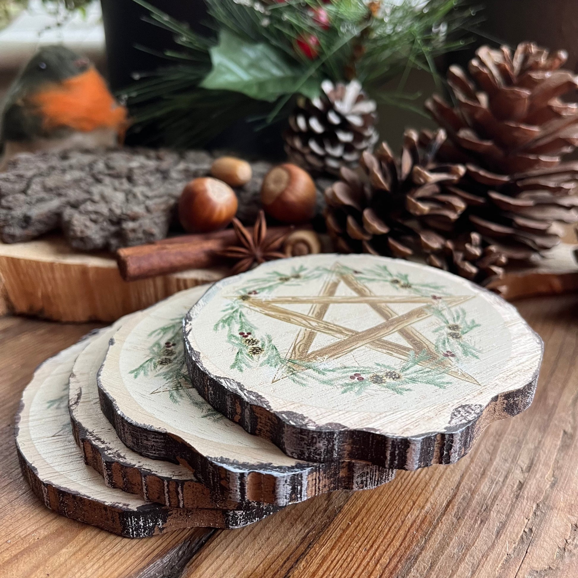 A set of 4 natural wood slice coasters featuring a pretty Winter Solstice wreath and pentagram design. Shown on a wooden table with natural seasonal symbols in the background such as pine cones, nuts and seeds and holly 