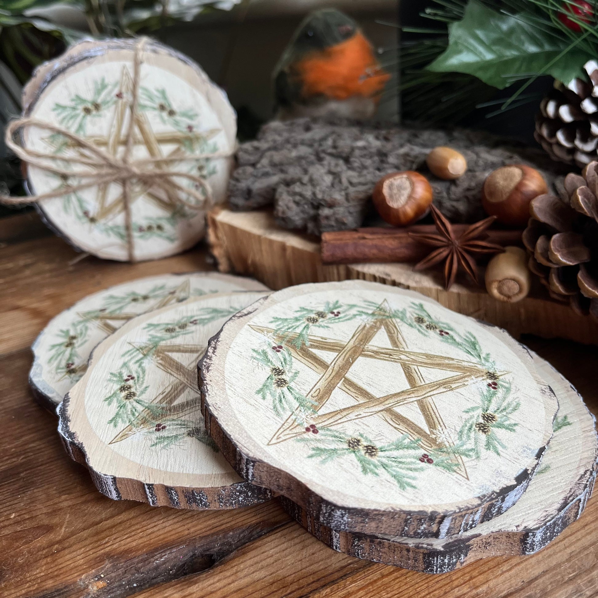 A set of 4 natural wood slice coasters featuring a pretty Winter Solstice wreath and pentagram design. Shown on a wooden table with natural seasonal symbols in the background such as pine cones, nuts and seeds and holly 