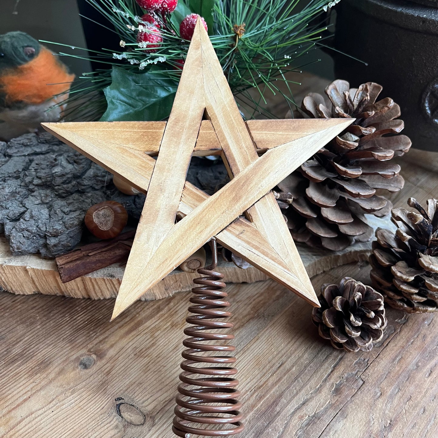 natural wooden Tree Topper in the shape of a pentagram star, shown with traditional christmas decorations of pine cones, holly and robin in the background 