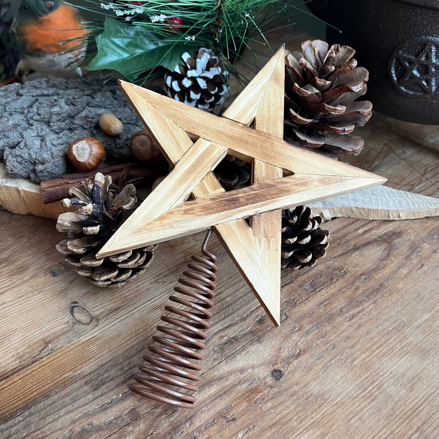 natural wooden Tree Topper in the shape of a pentagram star, shown with traditional christmas decorations of pine cones, holly and robin in the background 