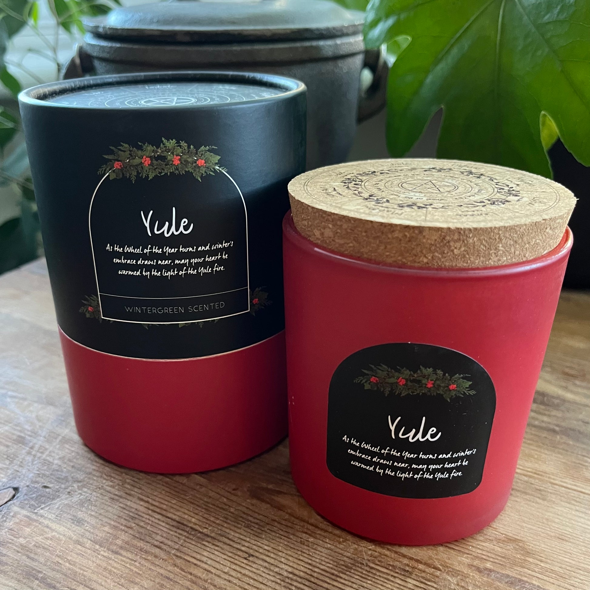 Two candles labeled 'Yule' on a wooden surface with plants in the background.