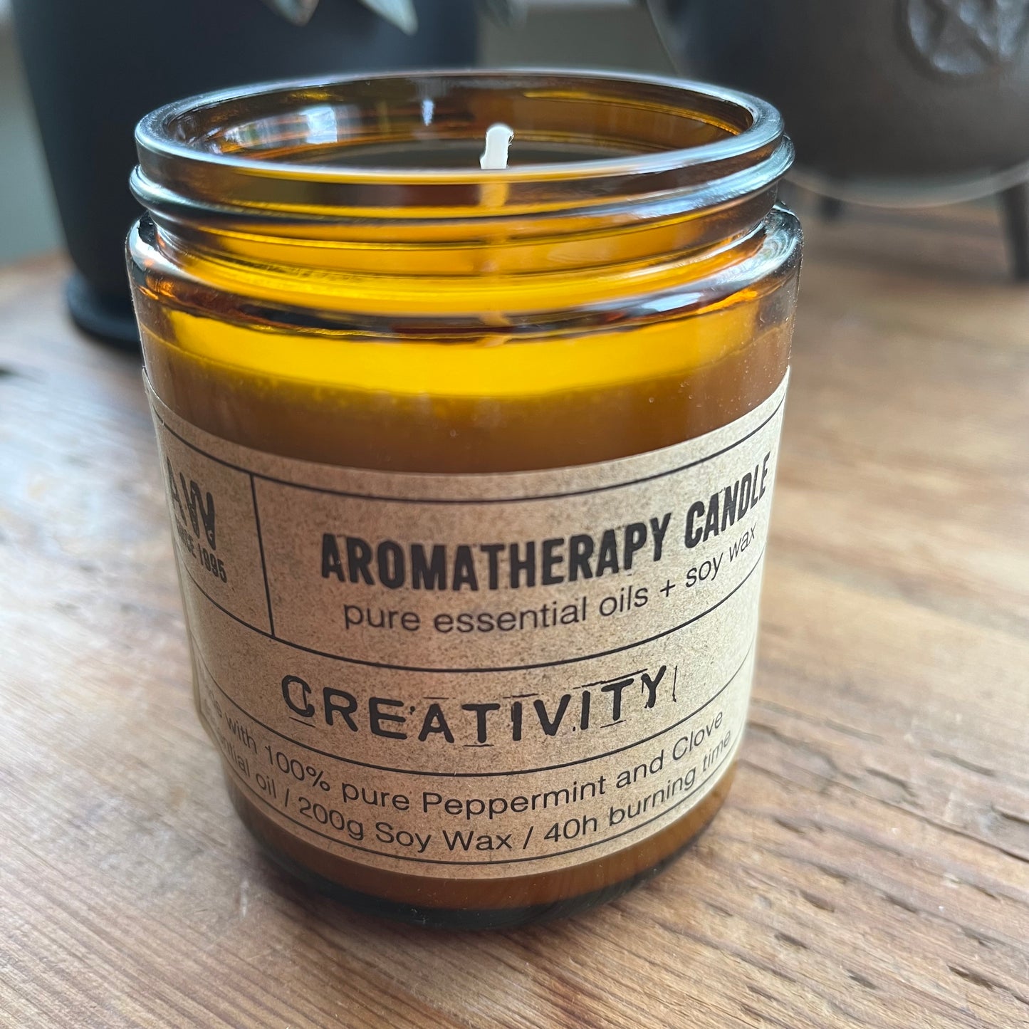 An amber glass jar candle with a beige paper label reading 'AROMATHERAPY CANDLE' and 'GREATIVITY!' on a wooden tabletop.