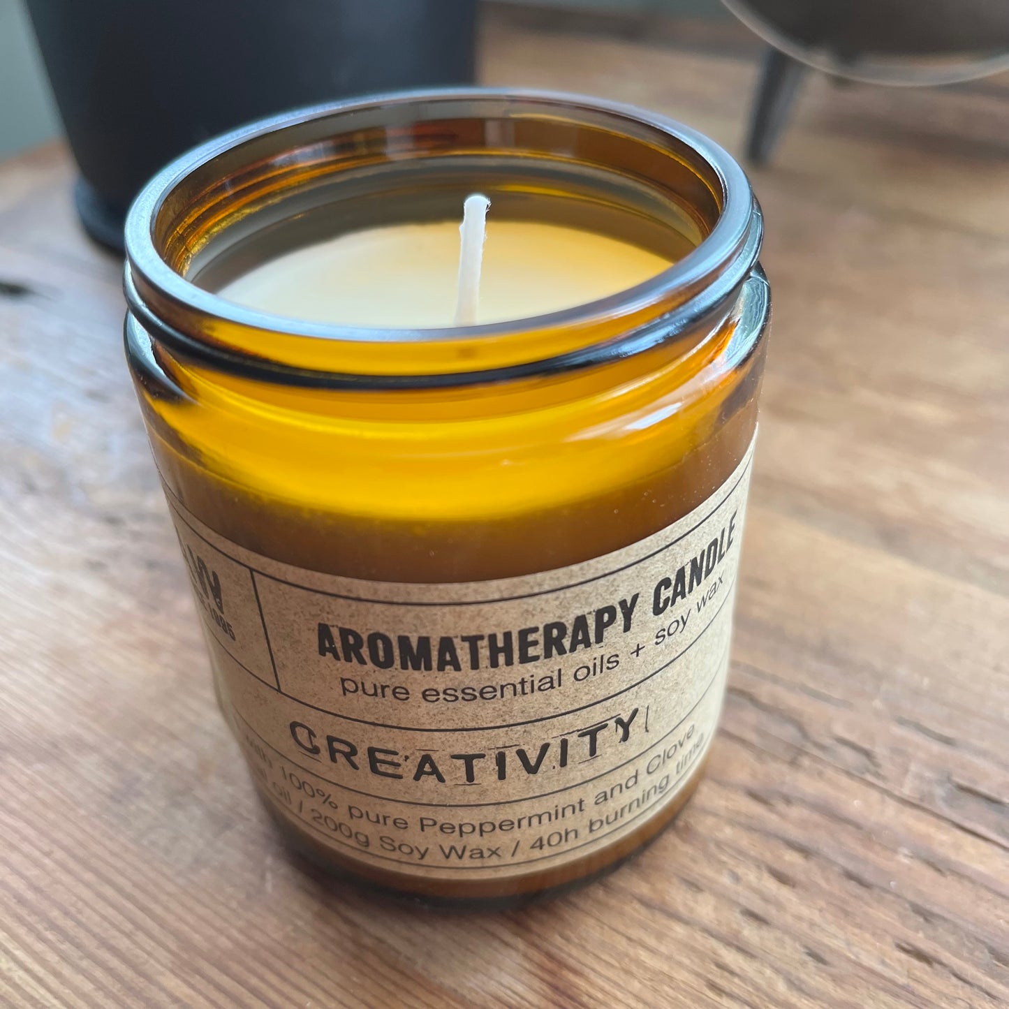 An amber glass jar candle labeled 'Aromatherapy Candle Creativity!' sits on a wooden tabletop. The candle has a beige label and a white wick, with a blurred background showing a dark container and a metal object.