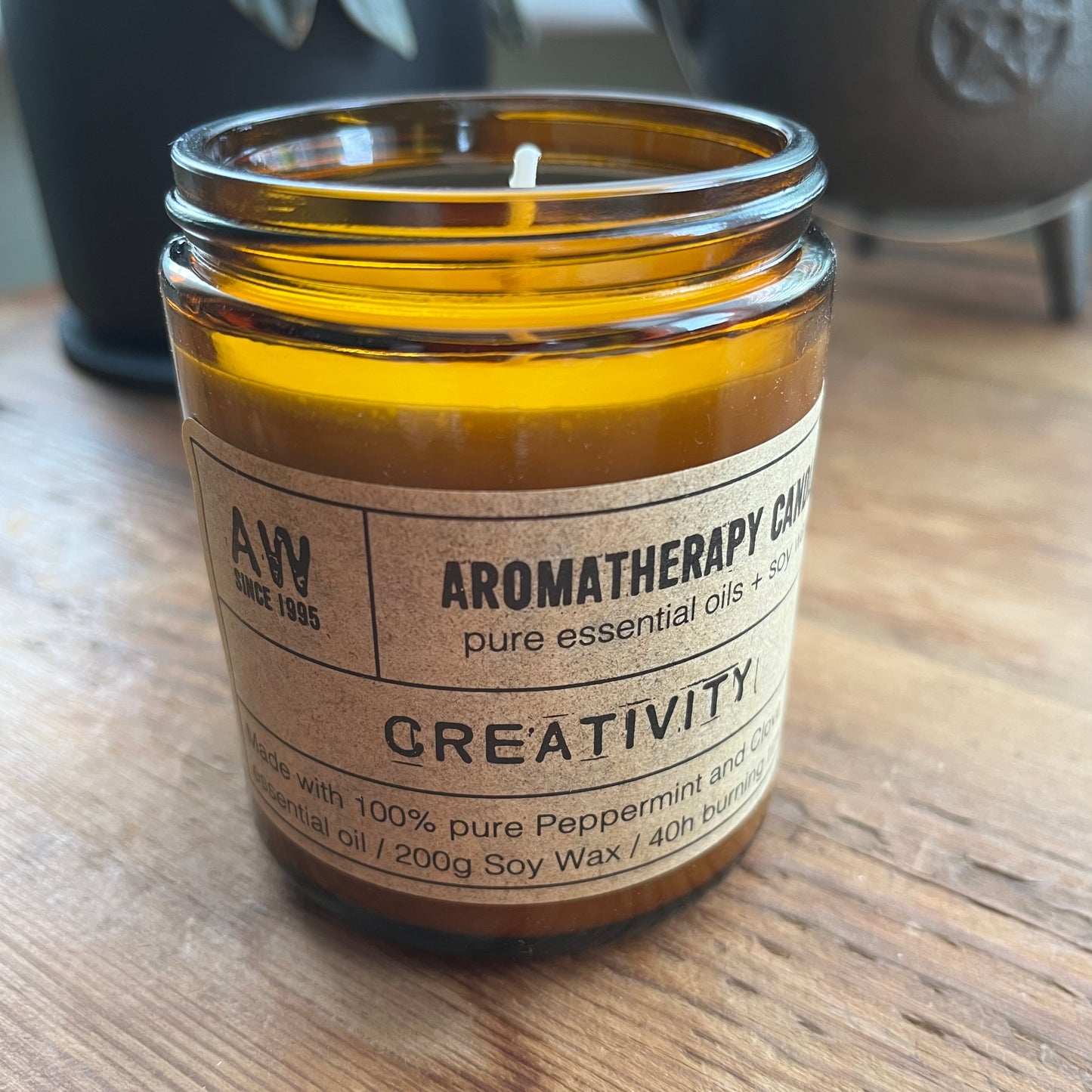A glass jar candle with amber-brown glass and a beige paper label reading 'Aromatherapy Candle' and 'Creativity', placed on a wooden surface.