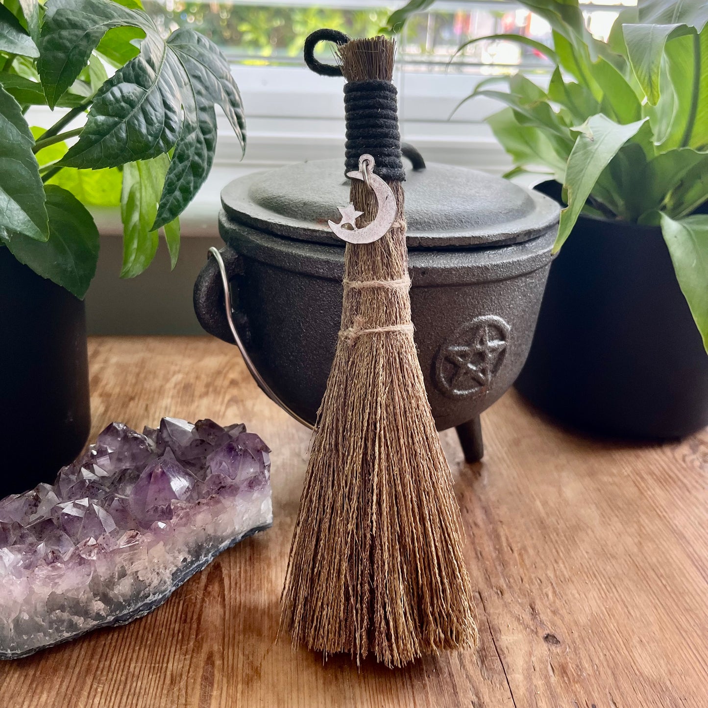 Small broom with a pentacle charm on a wooden surface next to a crystal and a cauldron.