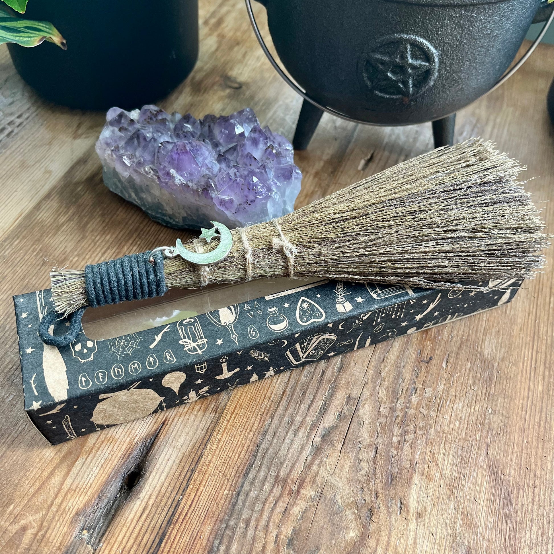 Broom with a handle on a decorative box, surrounded by crystals and a cauldron on a wooden surface.
