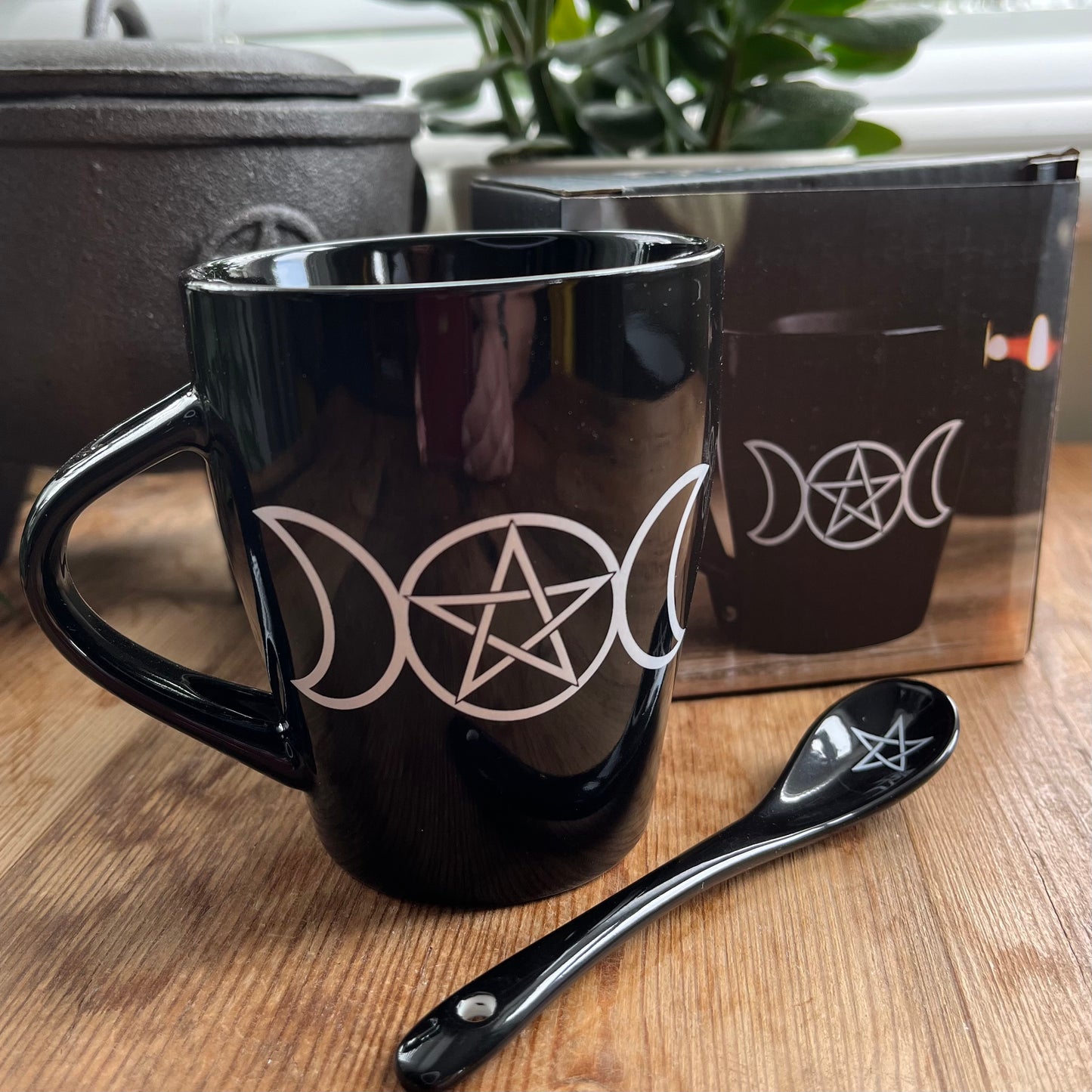 Black mug with a white triple moon and pentagram design on both sides. A matching ceramic poon slots into the handle of the mug 