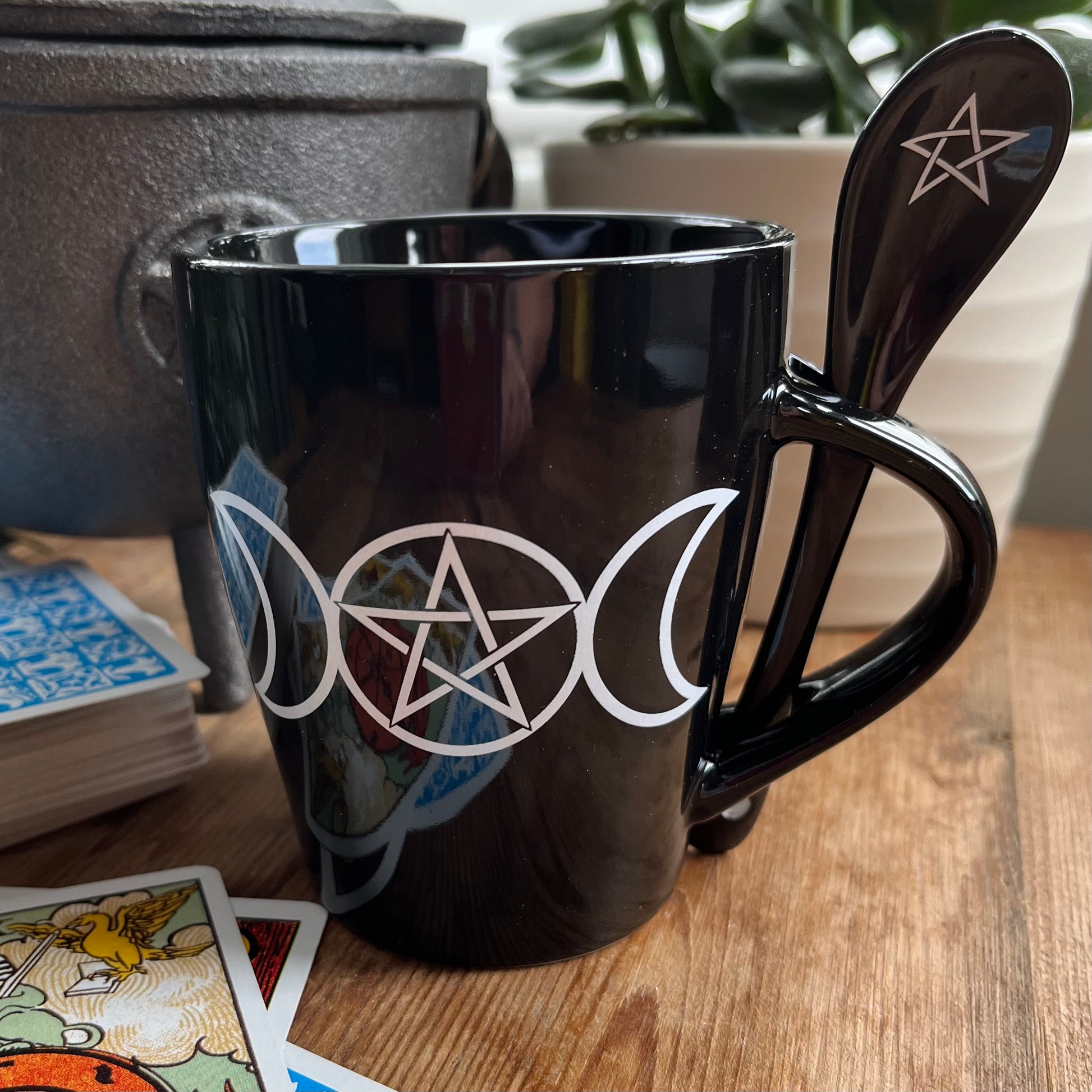 Black mug with a white triple moon and pentagram design on both sides. A matching ceramic poon slots into the handle of the mug 