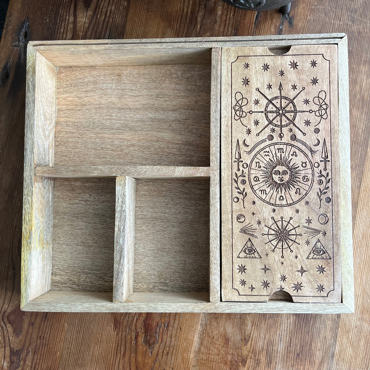 A rectangular wooden box with a divided interior and a front drawer featuring engraved occult-style symbols, including a sun face, stars, triangles, and a wheel, photographed on a wooden tabletop.