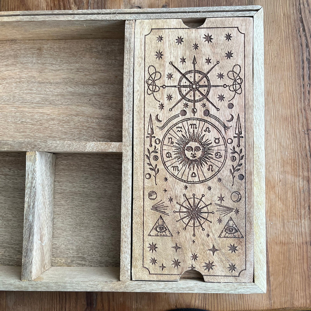 A wooden drawer or box with a front panel engraved with mystical symbols including a sun face, stars, triangles, and celestial motifs, shown on a wooden surface.