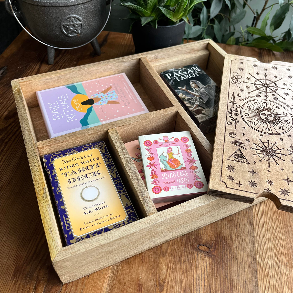 An open wooden divided tray holding several tarot card decks and a decorative wooden box lid with engraved celestial artwork, arranged on a wooden table with plants in the background.