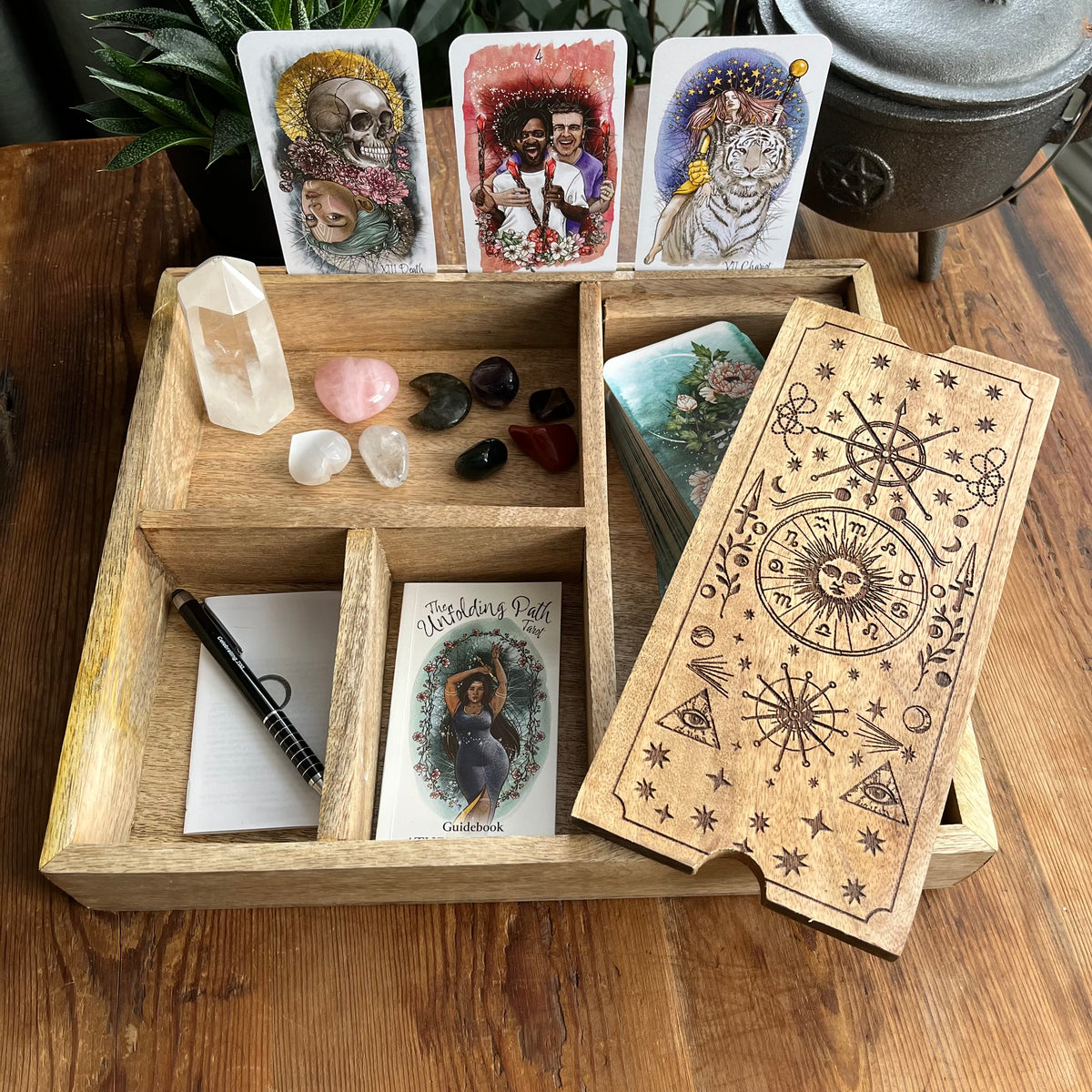 A wooden divided tray holding a tarot card deck, crystals, a notebook with a black pen, and printed cards, arranged on a wooden table with decorative plants and a metal vessel in the background.