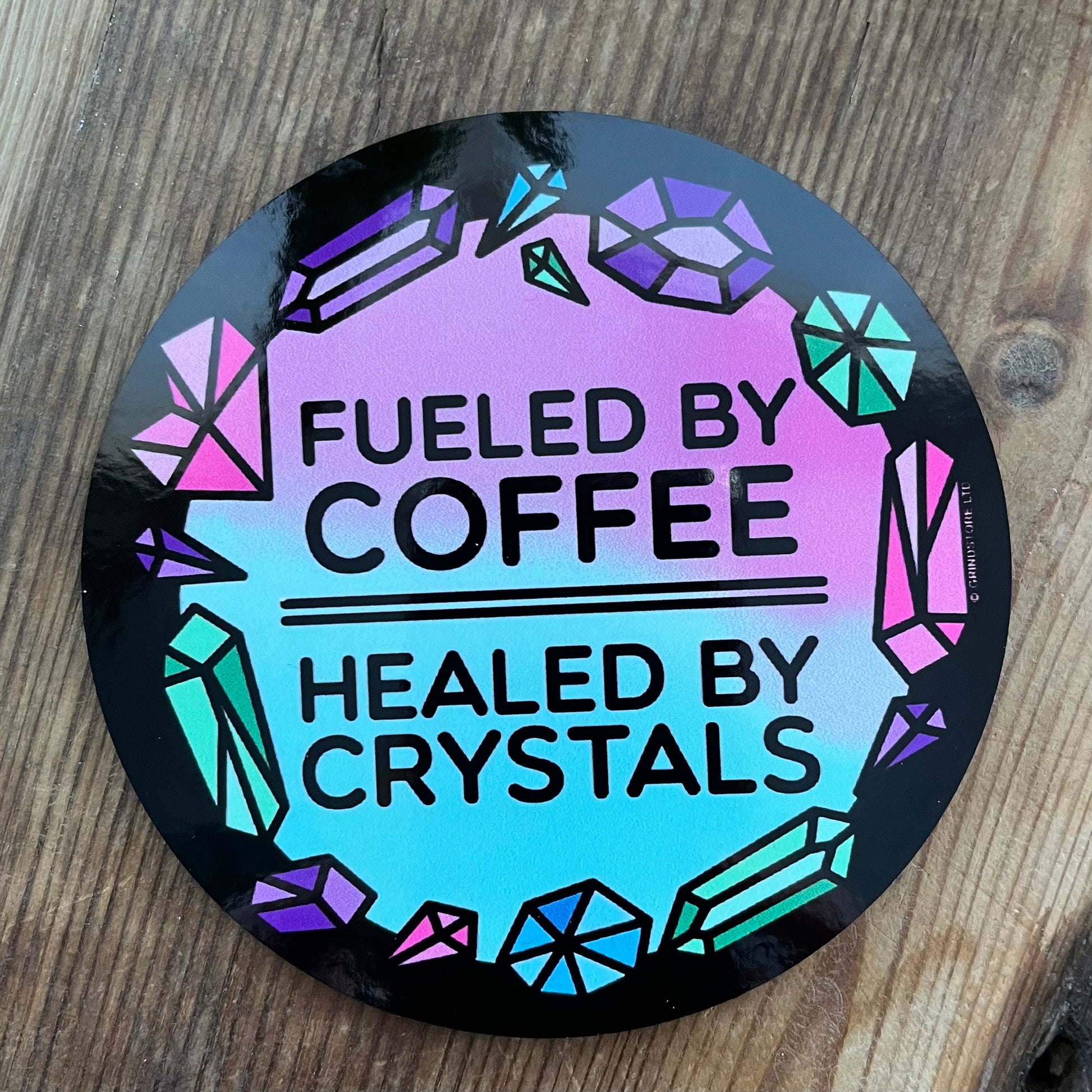 Round coaster with 'Fueled by Coffee, Healed by Crystals' text on a wooden surface