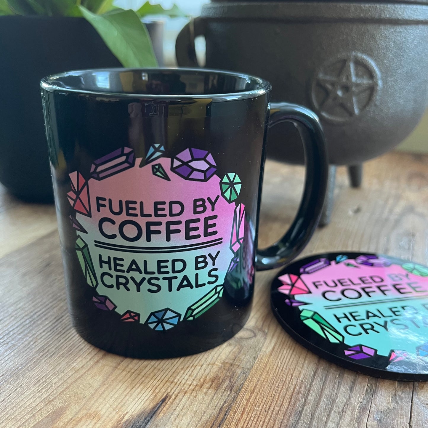 Black mug with colorful text 'Fueled by Coffee, Healed by Crystals' on a wooden surface.