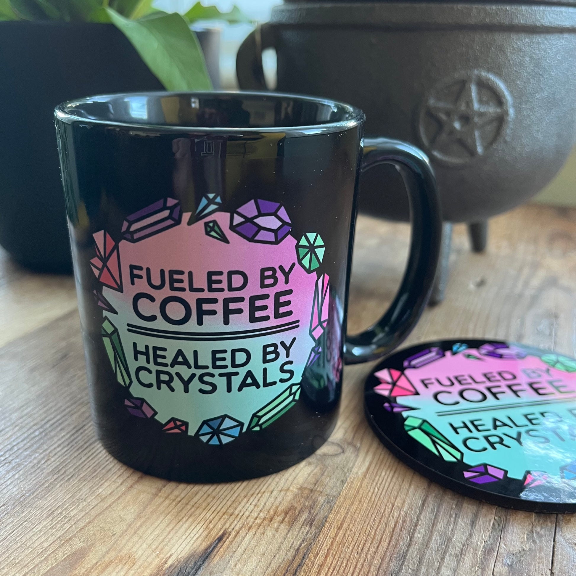 Black mug with colorful text 'Fueled by Coffee, Healed by Crystals' on a wooden surface.