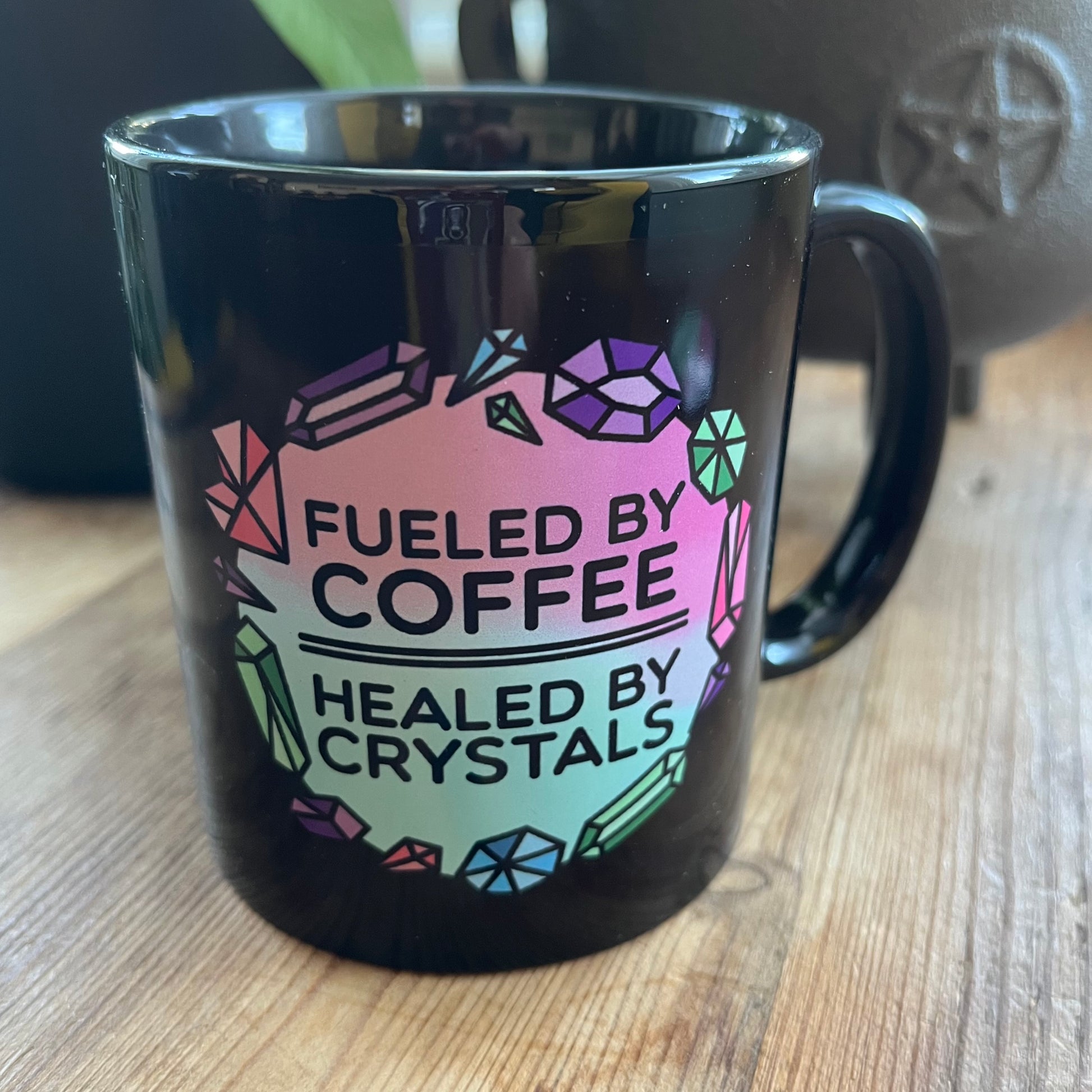 Black mug with colorful text 'Fueled by Coffee, Healed by Crystals' on a wooden surface.