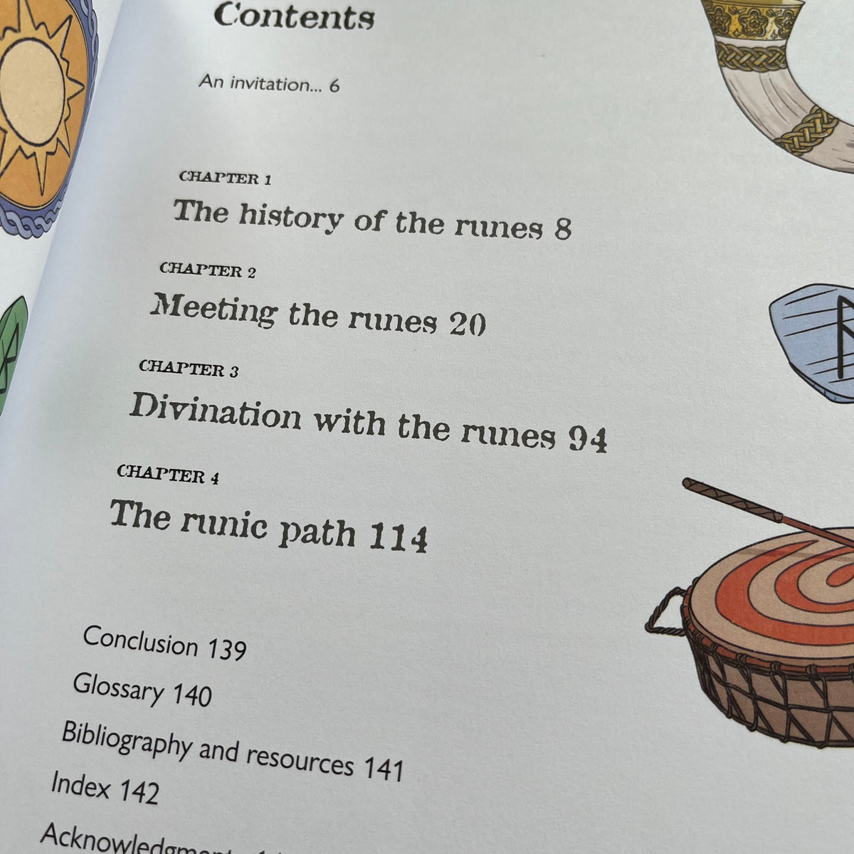 A close-up of an open book or booklet showing printed table-of-contents text, including chapter titles such as 'The history of the runes,' 'Meeting the runes,' 'Divination with the runes,' and 'The runic path.' Decorative illustrations of a sun, horn, shield, and drum appear along the right margin.