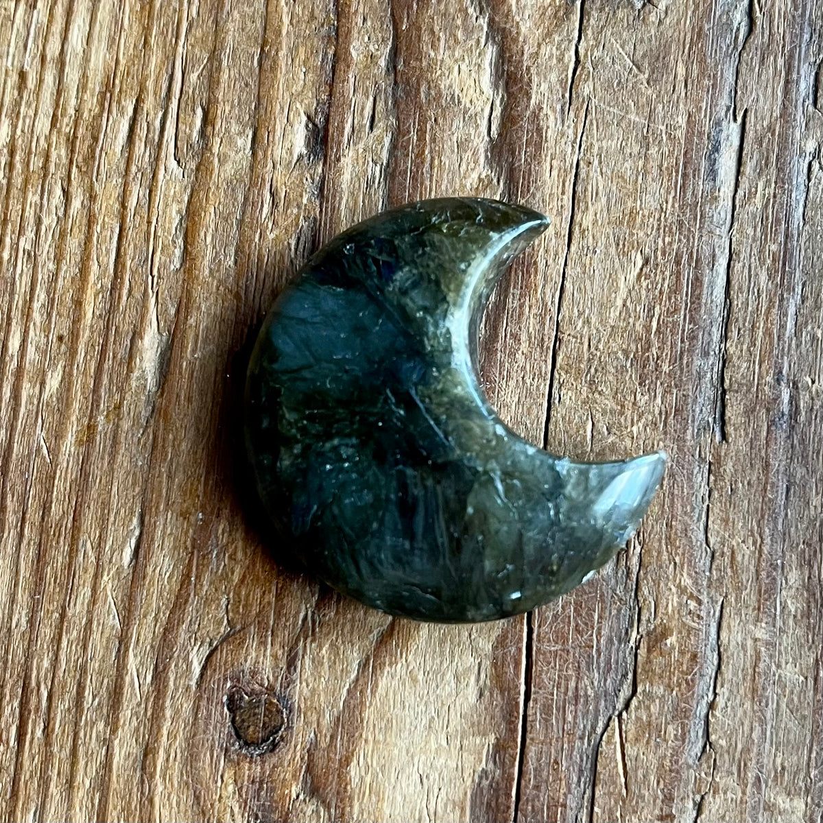 A polished crescent moon-shaped dark green stone charm resting on a rustic wooden surface.