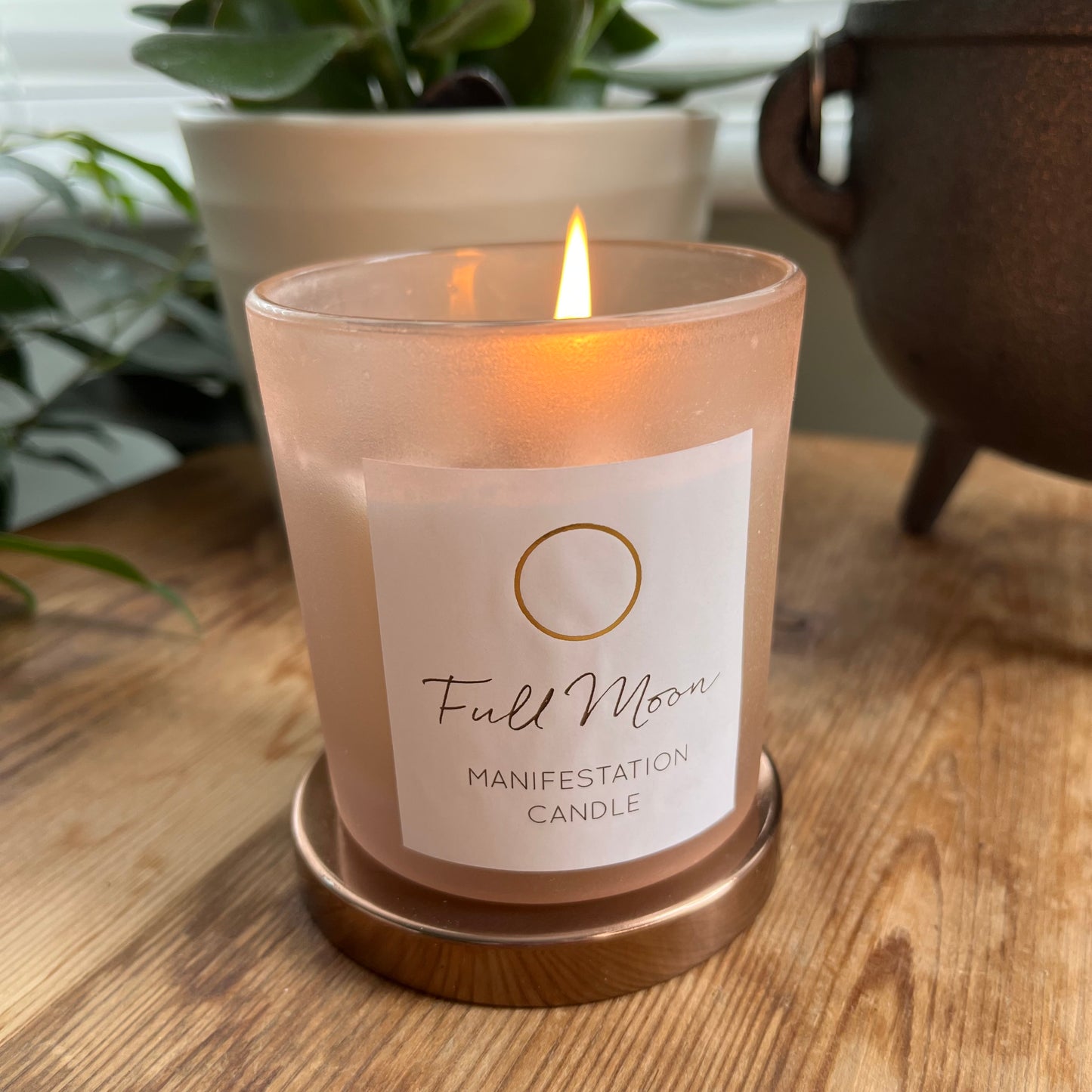 Candle contained within a beige frosted glass candle holder. the candle is sitting upon its bronze coloured metalic lid. the candle is labelled Full moon manifestation candle. there is a warm flame rising from the candle.