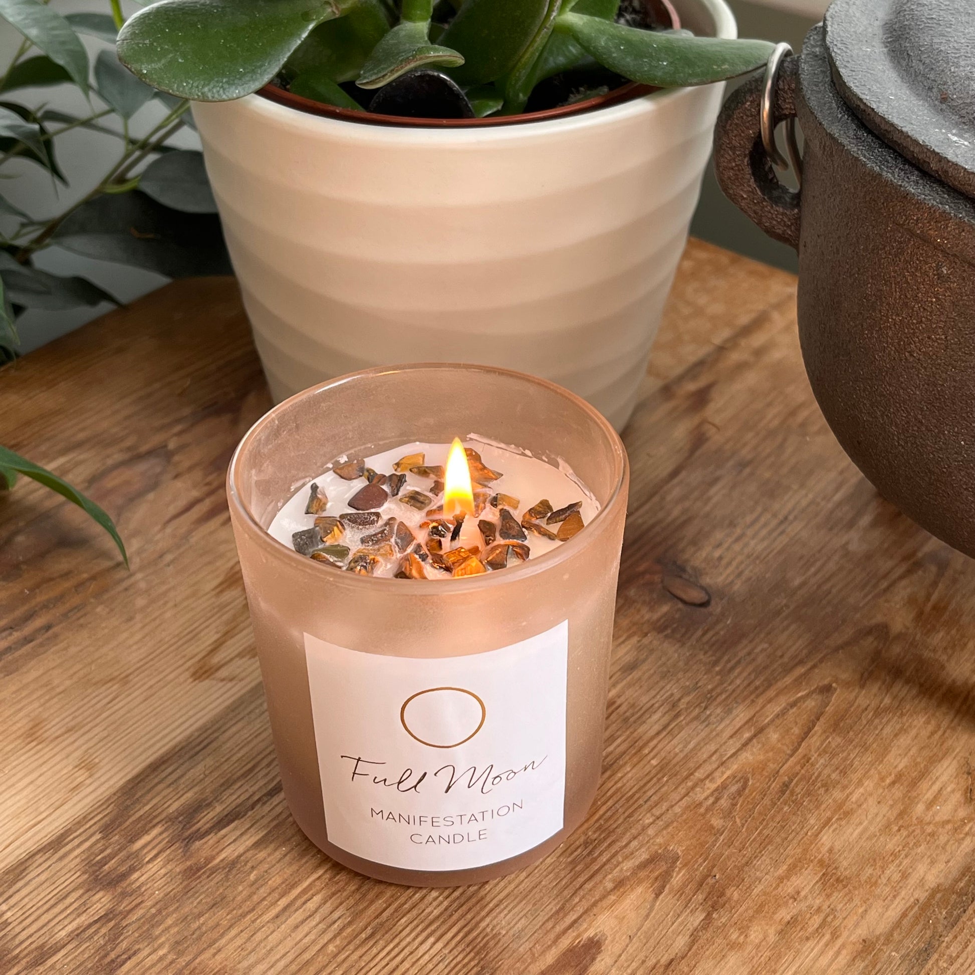 Beige frosted glass candle holder containing a white candle infused with Tigers eye crystals. Candle is labelled Full Moon Manifestation candle. the candle is lit and the candle flames creates a warm glow 