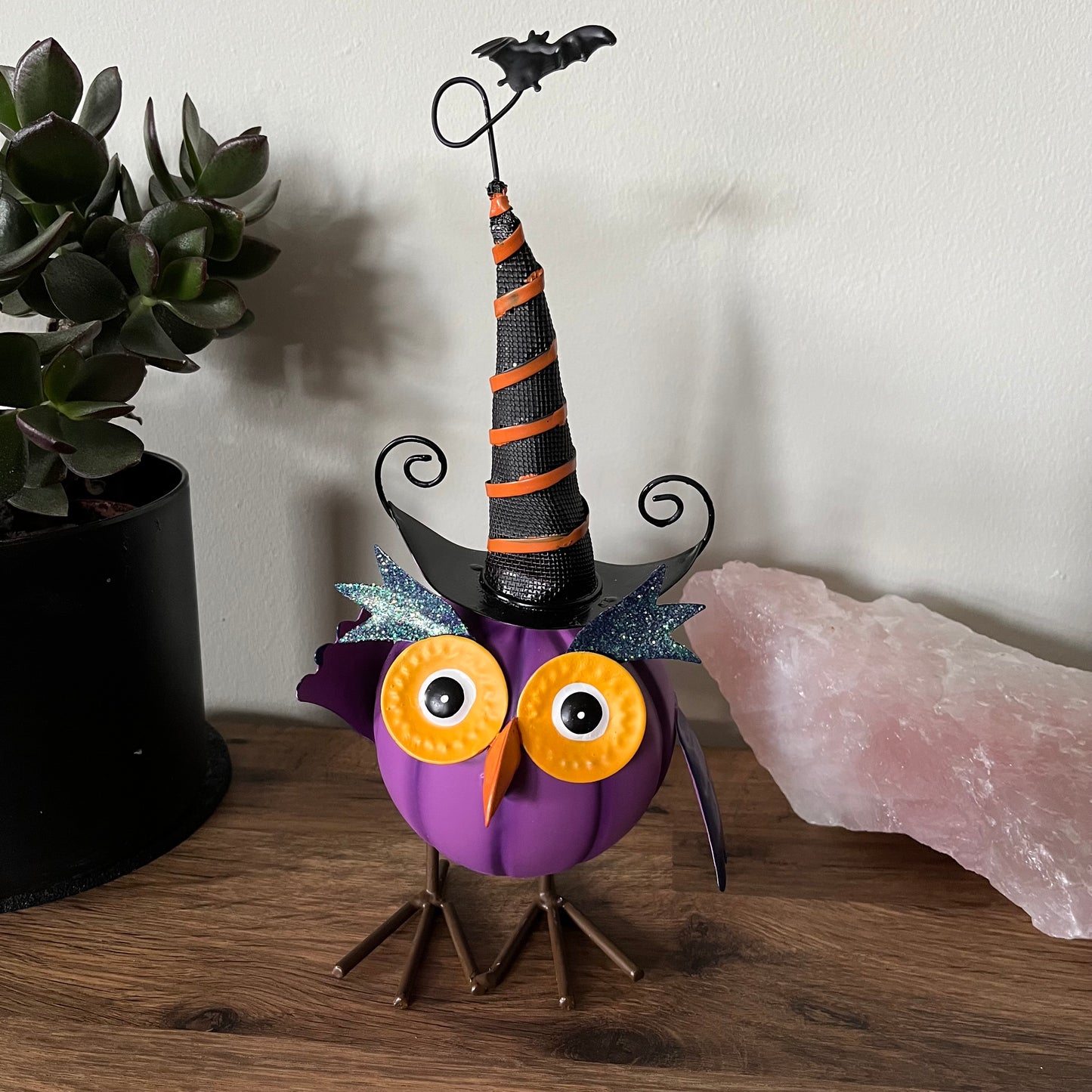 Halloween-themed owl figurine with a witch hat on a wooden surface next to a plant and crystal.
