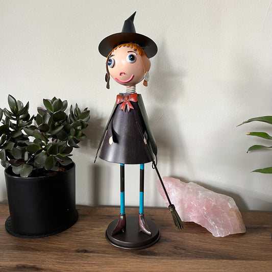 Decorative witch figurine with a broom, standing on a wooden surface next to a plant and a crystal.
