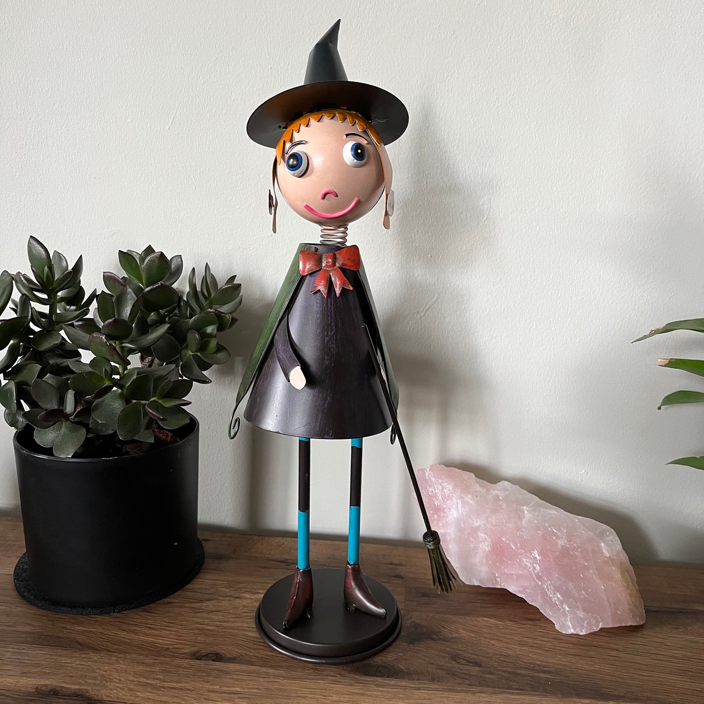 Decorative witch figurine with a broom next to a potted plant and crystal on a wooden surface.