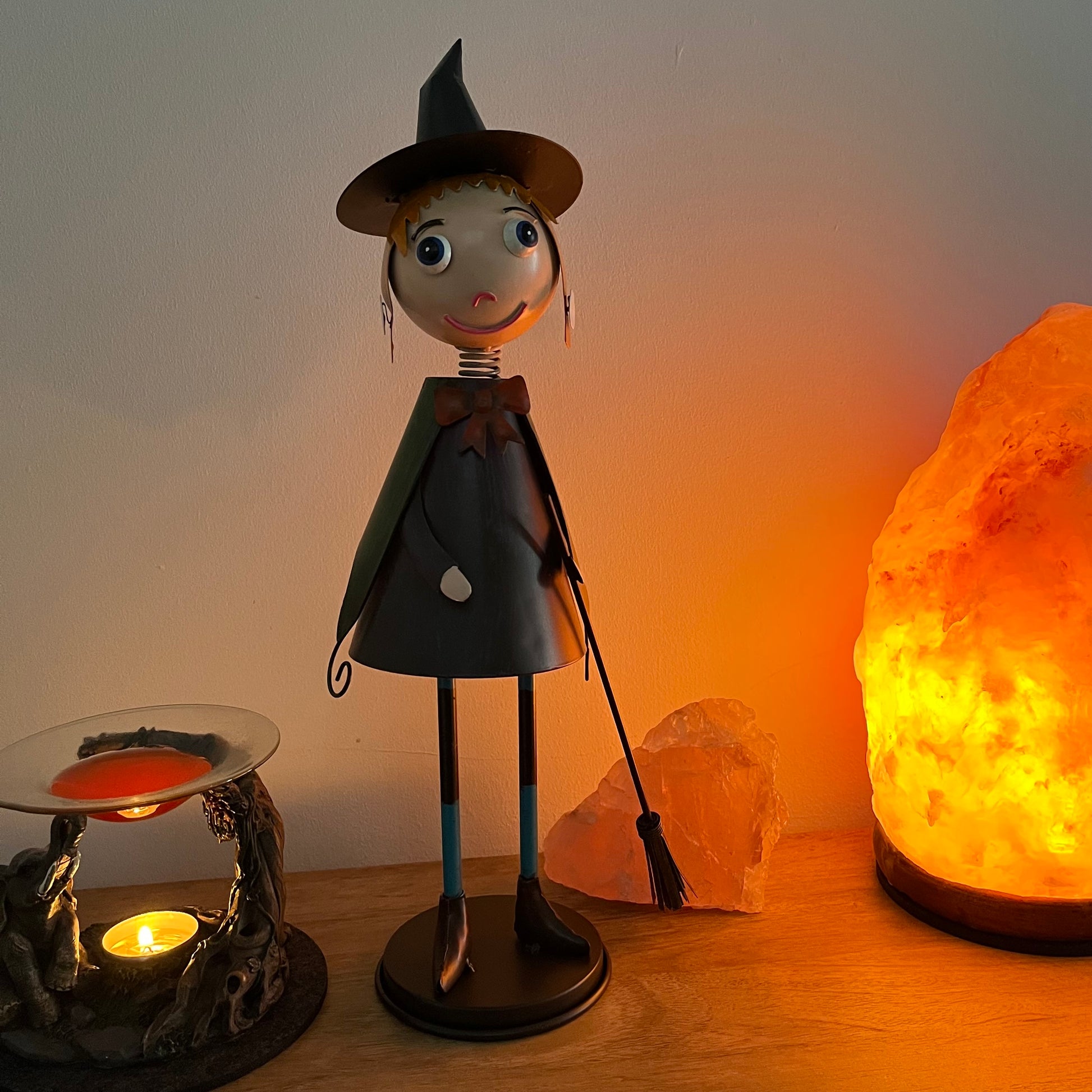Decorative witch figurine with a broom next to a salt lamp on a wooden surface.