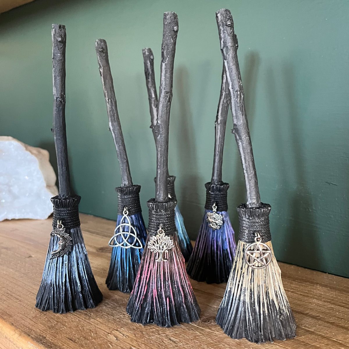 Six miniature decorative broomsticks with twig-style handles and black bristles are arranged on a wooden surface against a green wall. Each broom has a different coloured brush in shades of blue, purple, pink, beige, and dark gray, with small silver charms attached near the necks.