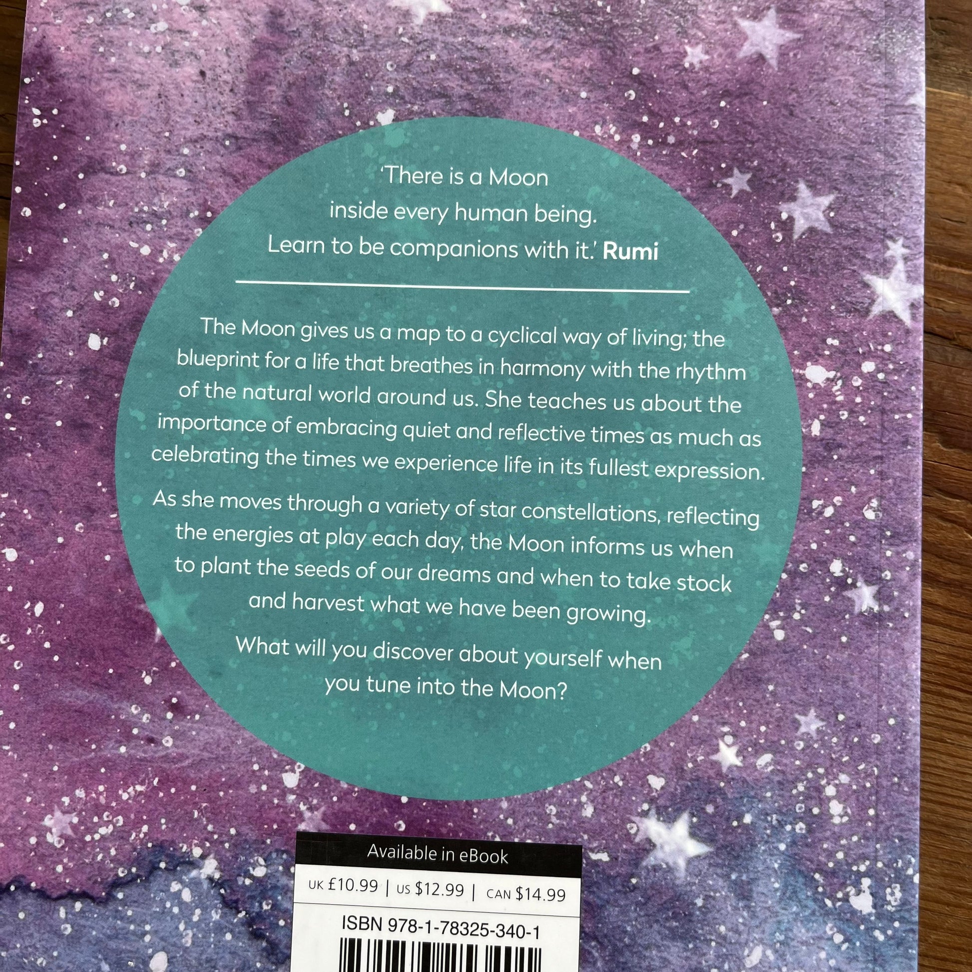 The back cover of a book titled 'Moon power' lies flat on a dark wooden table. The cover features a purple and blue galaxy watercolor design with white stars and a large teal circular text block in the center.