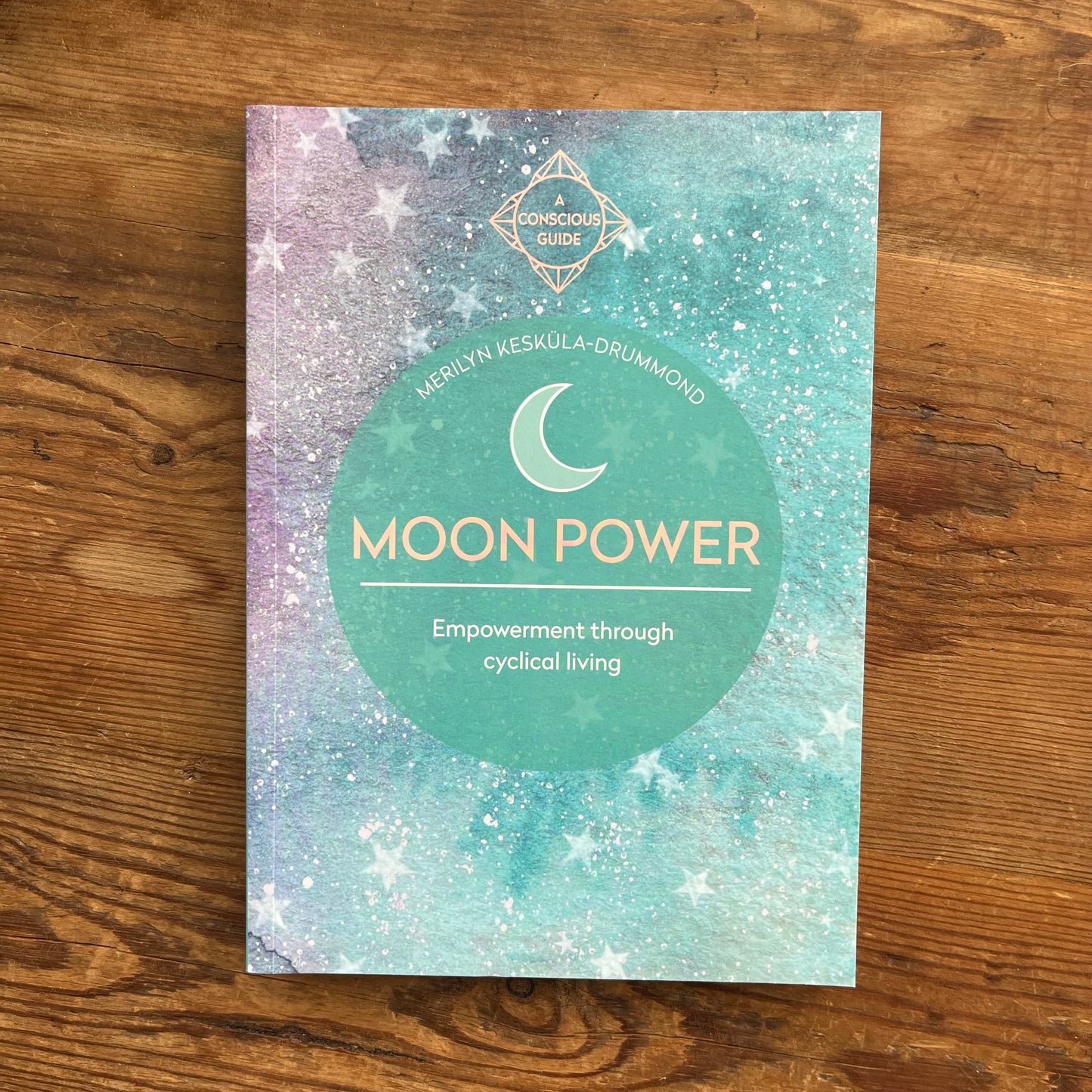 A book cover titled 'Moon Power' by Merilyn Keskula-Drummond, featuring a teal and lavender watercolor starry background with a large teal circle in the center, placed on a wooden surface.